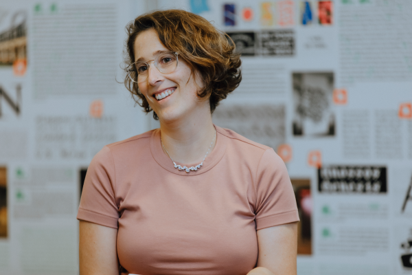 Portrait of Joana Correia smiling and looking to the side, pictured in front of a variety of documents and images on the wall.
